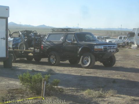 1990 Ford Bronco - black lifted Ford Bronco with a roof rack tows a trailer carrying an off-road buggy across a dusty desert lot.