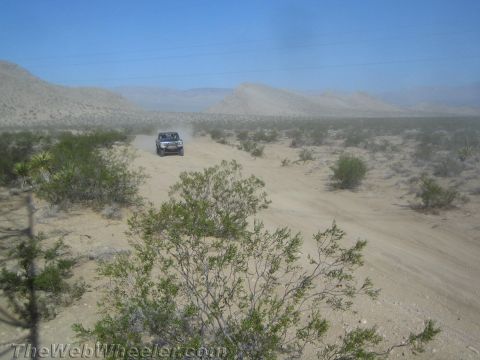 1990 Ford Bronco - small off-road truck drives along a sandy desert trail lined with brush, with mountains on the horizon.