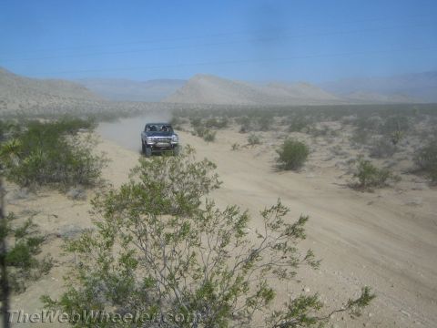 1990 Ford Bronco - light-colored off-road truck drives along a winding sandy desert trail with hills in the background.