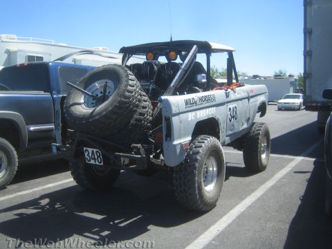1990 Ford Bronco - silver early Ford Bronco prerunner race truck numbered 348 with rear-mounted spare tire and roll cage, parked in a lot.