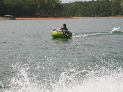 1985 Ford LTD Crown Victoria - A person enjoys tubing on a lake, with a wake trailing behind them and a treed shoreline in the distance.