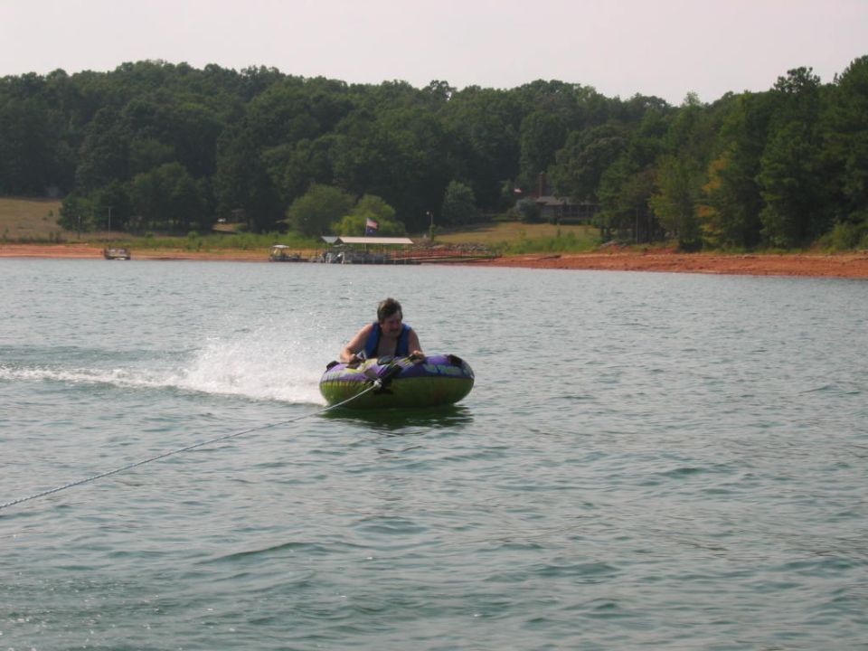 Person riding an inflatable tube behind a boat on a lake with trees and a dock in the background