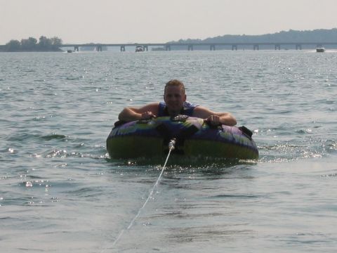 1985 Ford LTD Crown Victoria - A man enjoys tubing on a lake, with a bridge and boats visible in the distance.