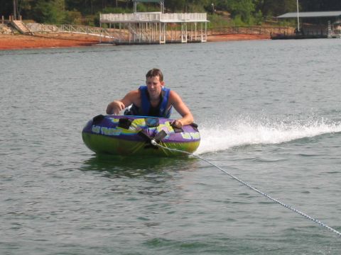 1985 Ford LTD Crown Victoria - A man enjoys tubing on a lake, with a dock and shoreline visible in the background.
