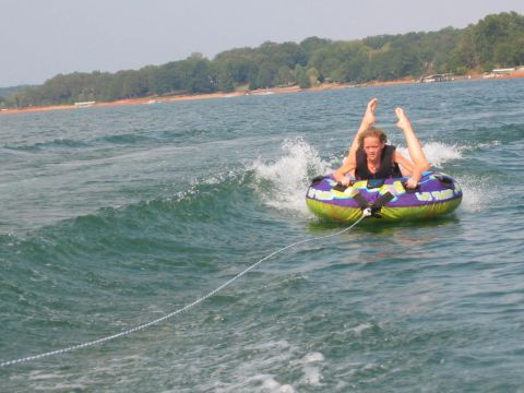 1985 Ford LTD Crown Victoria - A person enjoys tubing on a lake, with trees and a shoreline visible in the background.
