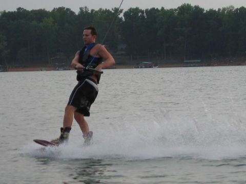 1985 Ford LTD Crown Victoria - A man wakeboards on a calm lake, creating a spray of water behind him as trees line the distant shore.