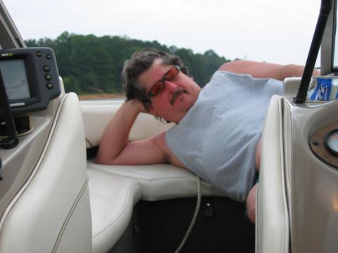 1985 Ford LTD Crown Victoria - A man relaxes on a boat on a lake, with trees in the background.