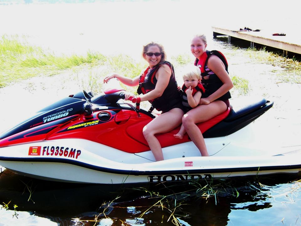 Red and black Honda AquaTrax jet ski with three riders in shallow water near a dock