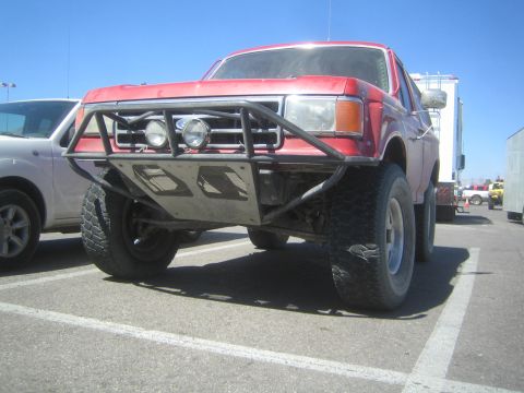 1990 Ford Bronco - Front quarter view of a lifted red full-size Ford Bronco with a black tube bumper and round driving lights on a sunny lot.