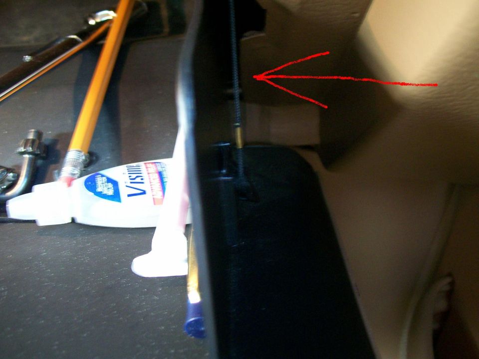 Close-up of a black plastic trim piece with a red arrow pointing to a cable, next to a tube of Visine and pencils