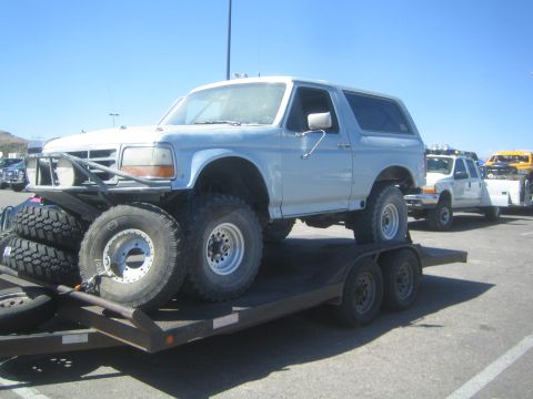 1990 Ford Bronco - lifted light-blue full-size Ford Bronco strapped on a flatbed trailer with spare tires, parked in a desert lot.