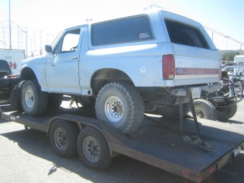 1990 Ford Bronco - Rear quarter view of a lifted light-blue full-size Ford Bronco with a FORD tailgate badge, strapped on a flatbed trailer.