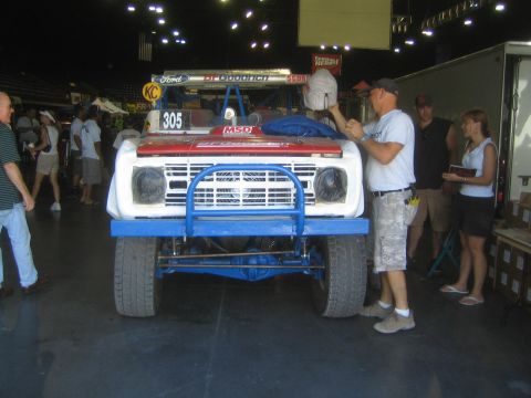 1990 Ford Bronco - Front view of a white and blue early Ford Bronco desert race truck numbered 305, with a man working on it indoors.