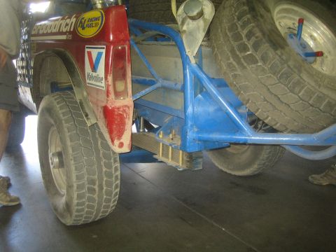 1990 Ford Bronco - Close-up of the front wheel, red fender and blue tubular bumper of an off-road race truck with sponsor decals.