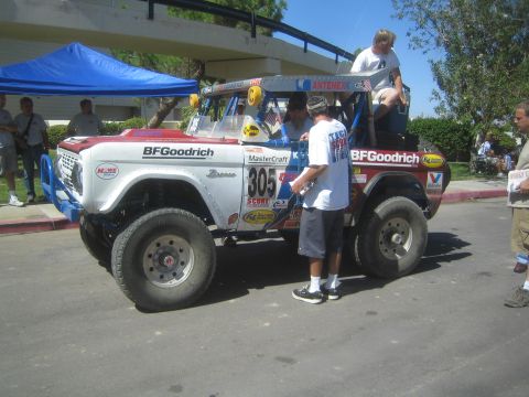 1990 Ford Bronco - lifted white early Ford Bronco off-road race truck numbered 805 with BFGoodrich livery parked outdoors as men work on it.