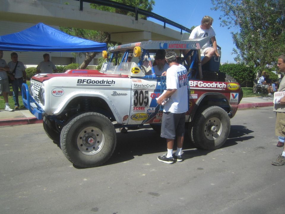 Lifted white and blue early Ford Bronco race truck number 805 parked outdoors with men around it