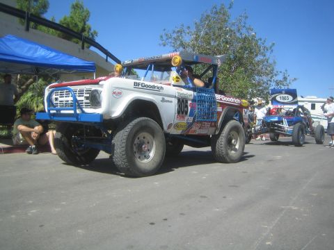 1990 Ford Bronco - White and blue early Ford Bronco off-road race truck numbered 305 parked on pavement with other race vehicles in the background.