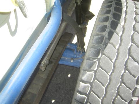 1990 Ford Bronco - Close-up of a blue tubular rocker rail, sliding mechanism and tire beside the white body panel of an off-road truck.