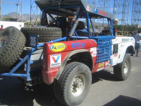 1990 Ford Bronco - Rear three-quarter view of a white and red early Ford Bronco off-road race truck numbered 805 with spare tires and a roll cage.