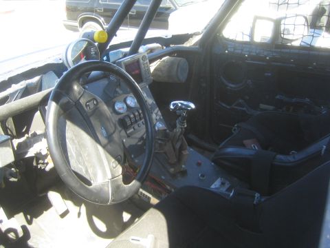 1990 Ford Bronco - Stripped race truck interior showing a Ford steering wheel, gauge cluster, shifter and a black bucket seat.