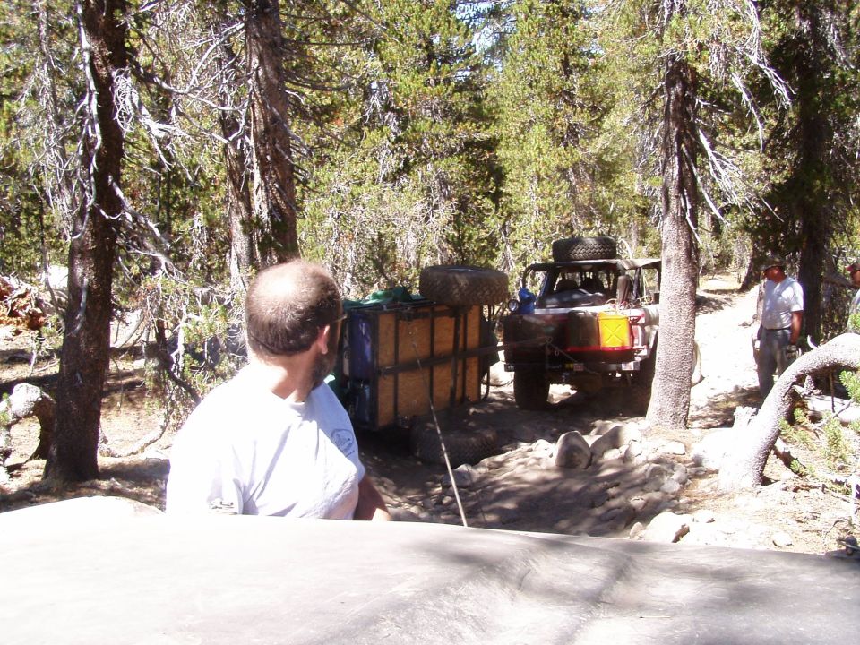 Open-top 4x4 towing loaded off-road trailer over rocks on a dusty pine forest trail