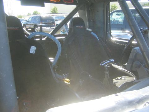 1990 Ford Bronco - Black MasterCraft bucket seats and roll cage of a race truck seen through the side window, with parked vehicles in the background.