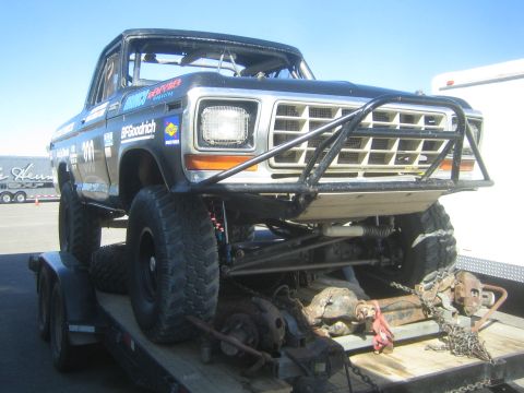 1990 Ford Bronco - black-and-orange older Ford off-road race truck numbered 300 sits on a flatbed trailer with spare parts under a clear blue sky.