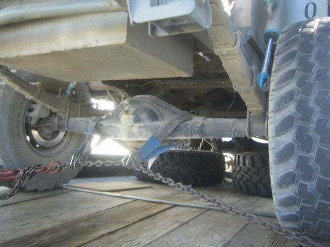 1990 Ford Bronco - Low-angle undercarriage shot of an off-road truck's rear axle, mud-terrain tires and tie-down chains on a wooden trailer deck.