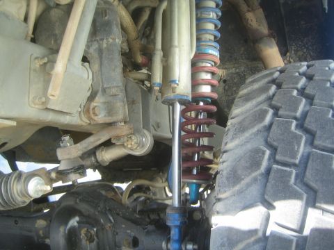 1990 Ford Bronco - Off-road truck front suspension detail showing a blue-and-red coilover shock, control arms, tie rod and a large tire.