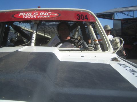 1990 Ford Bronco - driver sits behind the wheel inside a white off-road race truck cab marked 304, with a PHILS INC.com sticker on the roll cage.