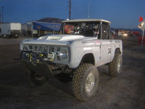 1990 Ford Bronco - lifted white early-Bronco-style off-road race truck numbered 348 sits on a dirt lot at dusk with trailers behind it.
