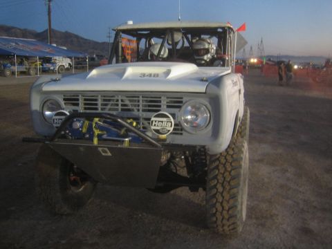 1990 Ford Bronco - Front three-quarter view of a white early-Bronco-style off-road race truck number 348 with Hella driving lights, parked on dirt at dusk.
