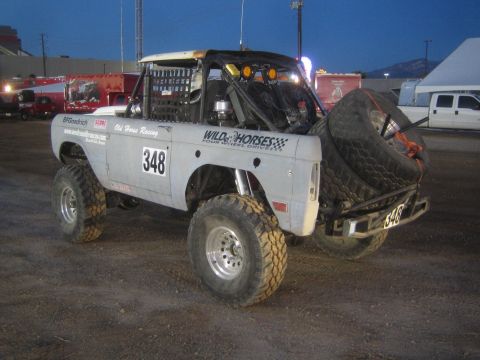 1990 Ford Bronco - white early-Bronco-style off-road race truck number 348 with a rear-mounted spare and Wild Horses Racing graphics, on dirt at dusk.