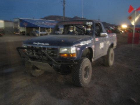 1990 Ford Bronco - dark blue lifted off-road race truck numbered 349 with a tube front bumper sits on a dirt lot at dusk.