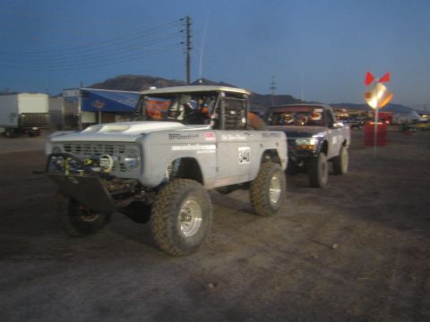 1990 Ford Bronco - white early-Bronco-style race truck numbered 348 stands beside a blue race truck 349 on a dirt staging lot at dusk.