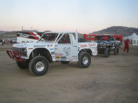 1990 Ford Bronco - white Ford off-road race truck numbered 2 with BFGoodrich and Wild Horses graphics is staged on a desert dirt lot with other trucks.
