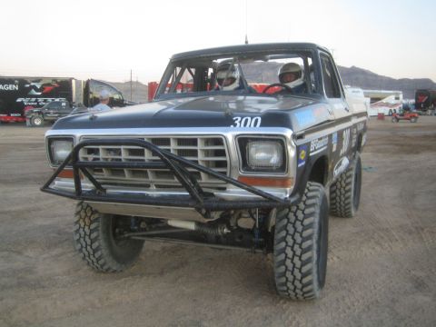 1990 Ford Bronco - dark blue late-1970s Ford pickup race truck numbered 300 with a tube front bumper and two helmeted occupants, on a dirt lot.