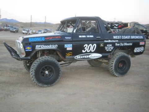 1990 Ford Bronco - black late-1970s Ford race truck numbered 300 with West Coast Broncos and Deaver Suspension graphics, parked on a dirt lot.