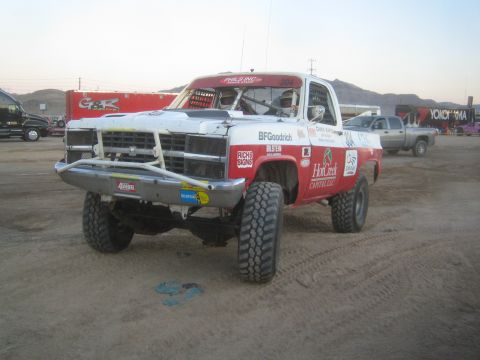 1990 Ford Bronco - white and red Chevrolet square-body off-road race truck numbered 304 with BFGoodrich graphics, on a desert dirt lot at dusk.