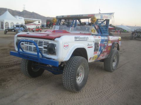 1990 Ford Bronco - white, red and blue early-Bronco off-road race truck numbered 305 with BFGoodrich graphics sits on a dirt lot at dusk.