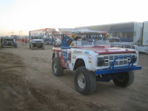 1990 Ford Bronco - red, white and blue early-Bronco race truck numbered 305 sits in a staging line beside support trailers at dawn.