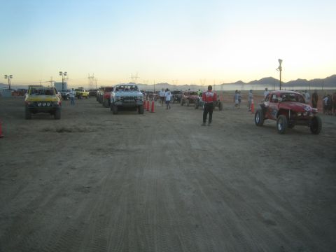 1990 Ford Bronco - Several off-road race trucks and a buggy are staged across a wide desert dirt lot with mountains and dusk sky behind.