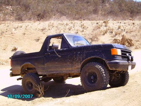 1990 Ford Bronco - A black Ford Bronco with large off-road tires is seen in a dry, hilly landscape.