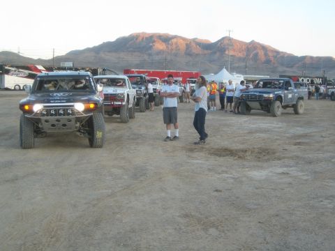 1990 Ford Bronco - black Toyota FJ Cruiser race vehicle leads a line of off-road trucks across a dirt lot as people stand nearby with mountains behind.