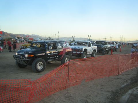 1990 Ford Bronco - black Toyota FJ off-road race truck numbered 2001 and other trucks line up behind an orange safety fence on a dirt lot at dawn.