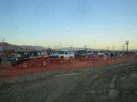 1990 Ford Bronco - long line of off-road race trucks stands behind an orange safety fence on a dirt lot with a crowd and power lines at dusk.