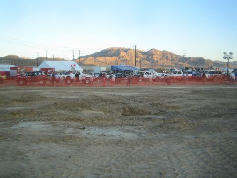 1990 Ford Bronco - distant row of off-road race trucks and a crowd gather behind red barriers on a churned dirt lot below rugged mountains.