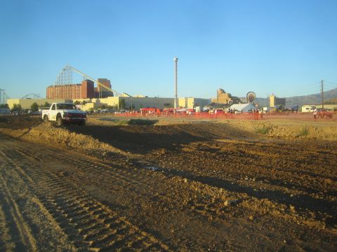 1990 Ford Bronco - white pickup truck drives along a dirt berm in the foreground with a fairground, roller coaster and crowd in the background.