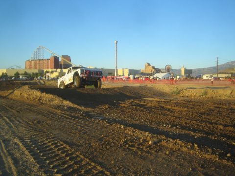 1990 Ford Bronco - white off-road race truck with a red front bumper drives along a dirt course with a fairground and roller coaster behind.