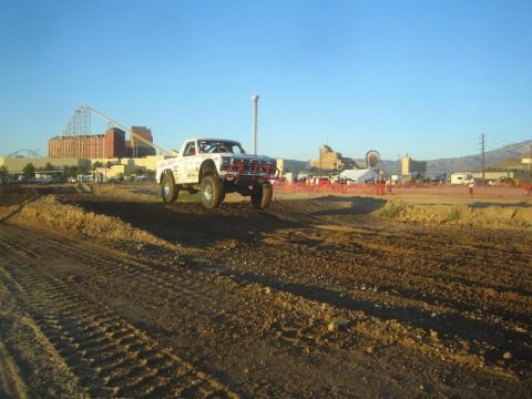 1990 Ford Bronco - white off-road race truck catches air over a dirt short-course track at golden hour, with a roller coaster and amusement park in the backgr…